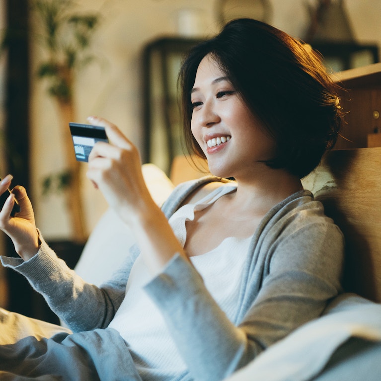 Beautiful smiling young Asian woman lying on the bed, shopping online on her smartphone and making mobile payment with credit card at home in the evening