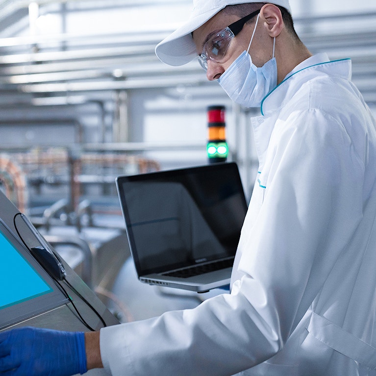 Man in a white robe and a mask with a laptop in his hands stands near the digital screen at the factory