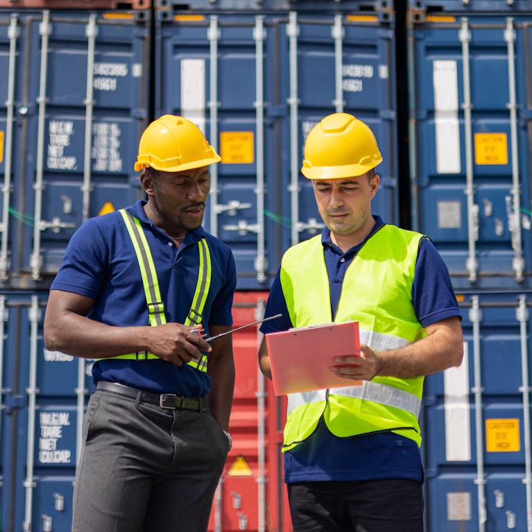 Two male coworkers wearing hardhats in front of shipping containers