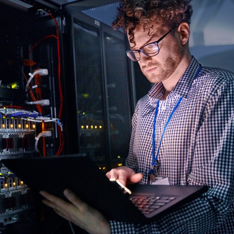 Focused male IT technician working at laptop in dark server room