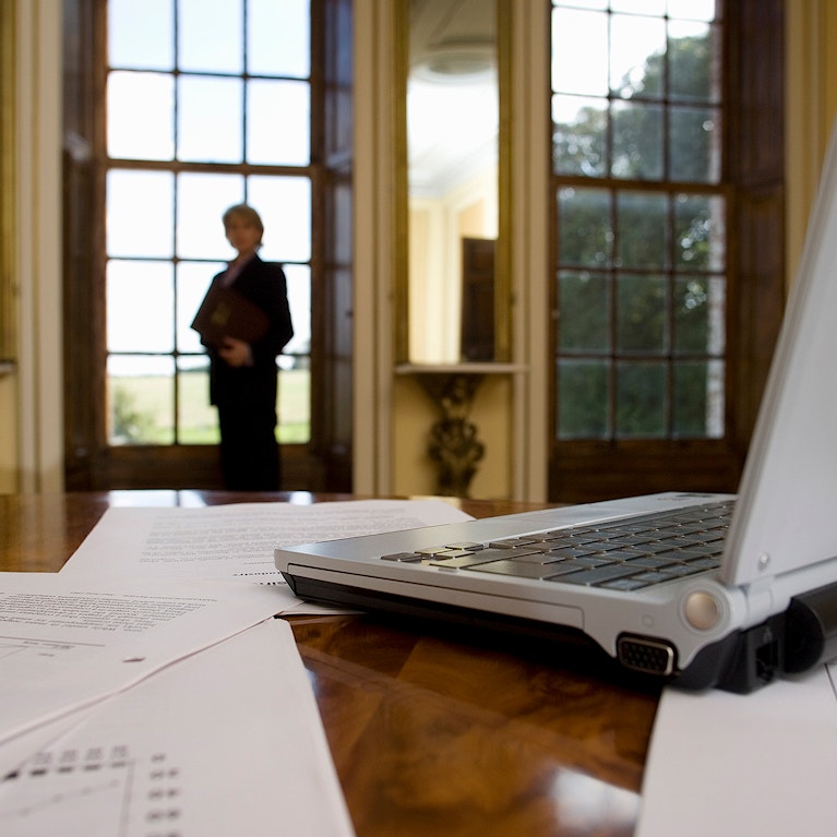 Laptop computer and paperwork on table, woman by window in background